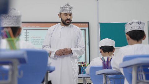An Omani Gulf Arab teacher wearing a dishdasha and kumma is explaining to his students in an advanced educational school environment, the modern education system, promoting sciences and innovations, and developing skills, monitoring a group of students inside the classroom, the quality of learning in academic schools.