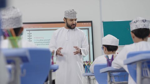 A group of students is being monitored inside the classroom, an Omani Gulf Arabic teacher wearing a dishdasha and kumma is explaining to his students in an advanced educational school environment, the modern education system, promoting sciences and innovations, and developing skills, the quality of learning in academic schools.