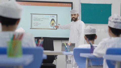 A group of students is being monitored inside the classroom, an Omani Gulf Arab teacher wearing a dishdasha and kumma is explaining to his students in an advanced educational school environment, the modern education system, promoting sciences and innovations, and developing skills, the quality of learning in academic schools.