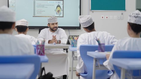 الاستفادة من التقنيات والتكنولوجيا الحديثة في التعلم والدراسة، طلاب عربيين خليجيين عمانيين يجلسون في مقاعدهم وأمامهم معلم يستخدم جهاز التابلت، المناهج المطورة والتعليم الالكتروني، تطور طرائق التدريس في مدارس سلطنة عمان