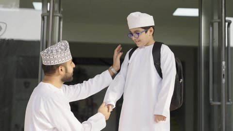 Academic schools in the Sultanate of Oman, an Arab Gulf Omani man wearing a dishdasha and kumma shakes hands with his son to enter the school lobby, gestures of happiness and joy, preparing for the new school semester.