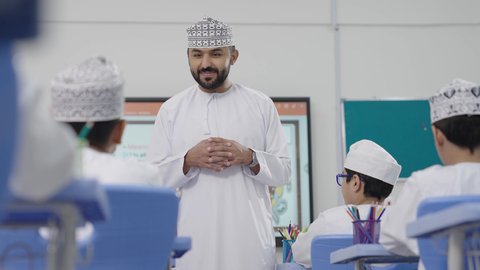 A group of students is being monitored inside the classroom, an Omani Gulf Arabic teacher wearing a dishdasha and kumma is explaining to his students in an advanced educational school environment, the modern education system, promoting sciences and innovations, and developing skills, the quality of learning in academic schools.