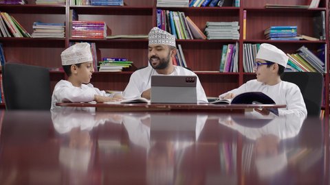 تطور طرائق التدريس ، المناهج المطورة والتعليم الالكتروني، الاستفادة من التقنيات والتكنولوجيا الحديثة في التعلم والدراسة،طلاب عربيين خليجيين عمانيين يرتدون الدشداشه والكمه يجلسون في المكتبة المدرسية مع معلم عماني يستخدم جهاز التابلت