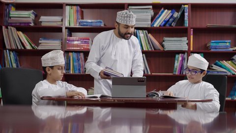 Two Gulf Arab Omani students wearing dishdasha and kumma are sitting in the school library with an Omani teacher using a tablet. The developed curricula and e-learning, benefiting from modern technologies and technology in learning and studying, and the evolution of teaching methods.