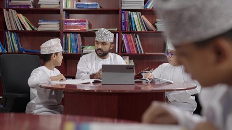 الاستفادة من التقنيات والتكنولوجيا الحديثة في التعلم والدراسة، المناهج المطورة والتعليم الالكتروني،تطور طرائق التدريس ، طلاب عربيين خليجيين عمانيين يرتدون الدشداشه والكمه يجلسون في المكتبة المدرسية مع معلم عماني يستخدم جهاز التابلت