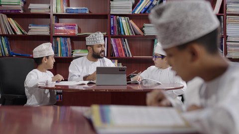 المناهج المطورة والتعليم الالكتروني، الاستفادة من التقنيات والتكنولوجيا الحديثة في التعلم والدراسة، تطور طرائق التدريس ، طلاب عربيين خليجيين عمانيين يرتدون الدشداشه والكمه يجلسون في المكتبة المدرسية مع معلم عماني يستخدم جهاز التابلت