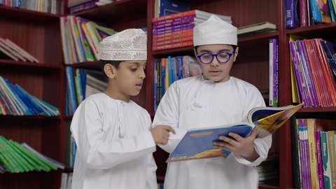 مفهوم التعليم والثقافه, بيئه مدرسيه تعليميه متطوره، الاستمتاع بقراءة الكتب المدرسية، طالبان   عربيان خليجيان عمانيان يرتديان الدشداشه والكمه احدهما يمسك الكتاب بيده فى المكتبه المدرسيه