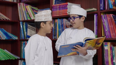 بيئه مدرسيه تعليميه متطوره، مفهوم التعليم والثقافه,الاستمتاع بقراءة الكتب المدرسية، طالبان   عربيان خليجيان عمانيان يرتديان الدشداشه والكمه احدهما يمسك الكتاب بيده فى المكتبه المدرسيه