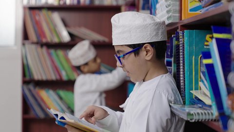 الاستمتاع بقراءة الكتب المدرسية، بيئه مدرسيه تعليميه متطوره، مفهوم التعليم والثقافه,ولد عربي خليجي عماني مبتسم يرتدي الدشداشه والكمه والحقيبة ونظارة يمسك الكتاب بيده فى المكتبه المدرسيه ، النظر الى الكاميرا بايماءات السرور والسعاده