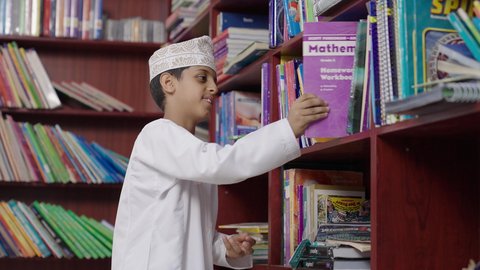 مفهوم التعليم والثقافه, بيئه مدرسيه تعليميه متطوره، الاستمتاع بقراءة الكتب المدرسية، ولد عربي خليجي عماني مبتسم يرتدي الدشداشه والكمه والحقيبة ونظارة يمسك الكتاب بيده فى المكتبه المدرسيه ، النظر الى الكاميرا بايماءات السرور والسعاده