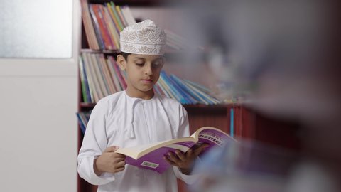 بيئه مدرسيه تعليميه متطوره، مفهوم التعليم والثقافه,الاستمتاع بقراءة الكتب المدرسية، ولد عربي خليجي عماني مبتسم يرتدي الدشداشه والكمه والحقيبة ونظارة يمسك الكتاب بيده فى المكتبه المدرسيه ، النظر الى الكاميرا بايماءات السرور والسعاده