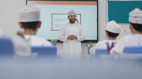 متابعة مجموعة من الطلاب داخل الفصل، معلم عربي عماني خليجي يرتدي الدشداشه والكمه يشرح لطلابه في بيئه مدرسيه تعليميه متطوره، نظام التعليم الحديث، تعزيز العلوم والإبتكارات وتنمية المهارات، جودة التعلم في المدارس الأكاديميه