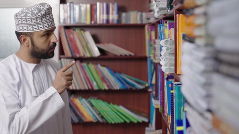 حب الاستطلاع والمعرفة ، بيئه مدرسيه تعليميه متطوره، مفهوم التعليم والثقافه,الاستمتاع بقراءة الكتب المدرسية، معلم  عربي خليجي عماني مبتسم يرتدي الدشداشه والكمه يمسك الكتاب بيده فى المكتبه المدرسيه