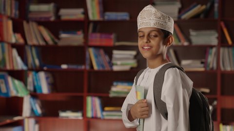 الاستمتاع بقراءة الكتب المدرسية، التعليم فى سلطنة عمان ،ولد عربي خليجي عماني مبتسم يرتدي الدشداشه والكمه والحقيبة يقف ويمسك الكتاب بيده فى المكتبه المدرسيه ، النظر الى الكاميرا بايماءات السرور والسعاده