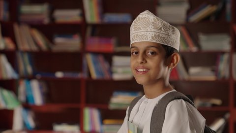 التعليم فى سلطنة عمان ، الاستمتاع بقراءة الكتب المدرسية، ولد عربي خليجي عماني مبتسم يرتدي الدشداشه والكمه والحقيبة يقف ويمسك الكتاب بيده فى المكتبه المدرسيه ، النظر الى الكاميرا بايماءات السرور والسعاده