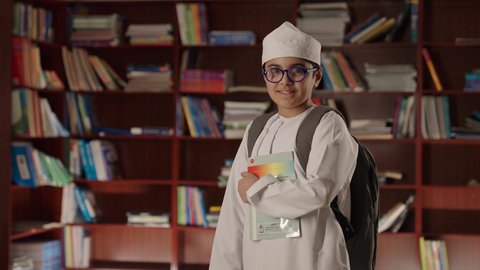 مفهوم التعليم والثقافه, بيئه مدرسيه تعليميه متطوره، الاستمتاع بقراءة الكتب المدرسية، ولد عربي خليجي عماني مبتسم يرتدي الدشداشه والكمه والحقيبة ونظارة طبية يقف ويمسك الكتاب بيده فى المكتبه المدرسيه ، النظر الى الكاميرا بايماءات السرور والسعاده