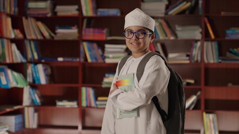 A developed educational school environment, the concept of education and culture, enjoying reading school books, a smiling Arab Gulf Omani boy wearing a dishdasha and kumma, holding a bag and wearing glasses, standing and holding a book in his hand in the school library, looking at the camera with gestures of joy and happiness.