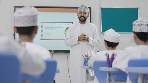 متابعة مجموعة من الطلاب داخل الفصل، معلم عربي عماني خليجي يرتدي الدشداشه والكمه يشرح لطلابه في بيئه مدرسيه تعليميه متطوره، نظام التعليم الحديث، تعزيز العلوم والإبتكارات وتنمية المهارات، جودة التعلم في المدارس الأكاديميه