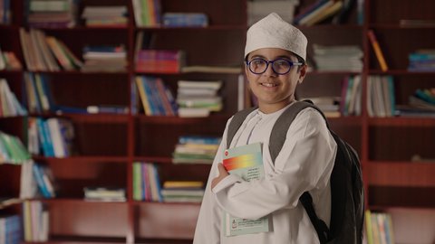 النظر الى الكاميرا بايماءات السرور والسعاده، بيئه مدرسيه تعليميه متطوره، مفهوم التعليم والثقافه,الاستمتاع بقراءة الكتب المدرسية، ولد عربي خليجي عماني مبتسم يرتدي الدشداشه والكمه والحقيبة ونظارة طبية يقف ويمسك الكتاب بيده فى المكتبه المدرسيه