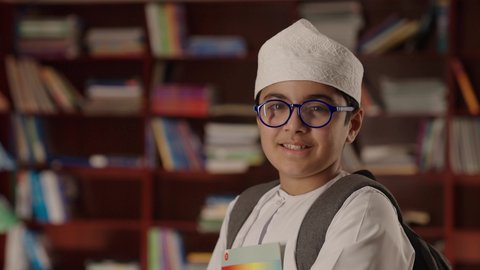 تنمية مهارة القراءة للطلاب, مفهوم التعليم والثقافه,الاستمتاع بقراءة الكتب المدرسية، ولد عربي خليجي عماني مبتسم يرتدي الدشداشه والكمه والحقيبة ونظارة طبية يقف ويمسك الكتاب بيده فى المكتبه المدرسيه ، النظر الى الكاميرا بايماءات السرور والسعاده