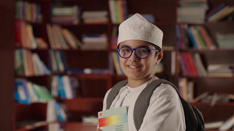 A developed educational school environment, the concept of education and culture, enjoying reading school books, a smiling Arab Gulf Omani boy wearing a dishdasha and kumma, holding a bag and wearing glasses, standing and holding a book in his hand in the school library, looking at the camera with gestures of joy and happiness.