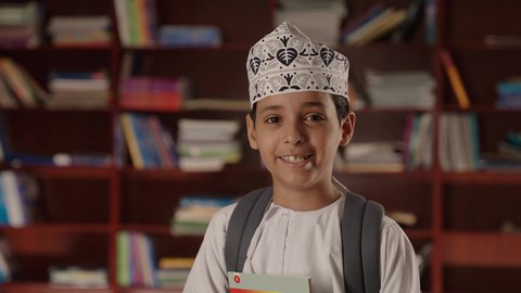 الاستمتاع بقراءة الكتب المدرسية، التعليم فى سلطنة عمان ،ولد عربي خليجي عماني مبتسم يرتدي الدشداشه والكمه والحقيبة يقف ويمسك الكتاب بيده فى المكتبه المدرسيه ، النظر الى الكاميرا بايماءات السرور والسعاده