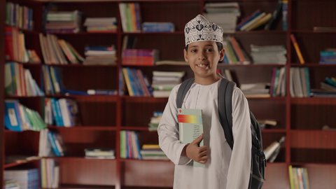 التعليم فى سلطنة عمان ، الاستمتاع بقراءة الكتب المدرسية، ولد عربي خليجي عماني مبتسم يرتدي الدشداشه والكمه والحقيبة يقف ويمسك الكتاب بيده فى المكتبه المدرسيه ، النظر الى الكاميرا بايماءات السرور والسعاده