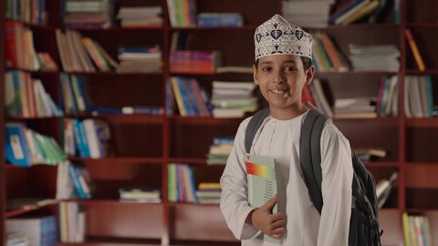 Expressions of joy and happiness, education in the Sultanate of Oman, enjoying reading school books, a smiling Arab Gulf Omani boy wearing a dishdasha and kumma, standing and holding a book in his hand in the school library.