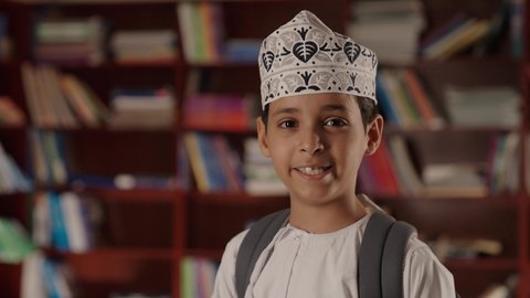 Education in the Sultanate of Oman, enjoying reading school books, a smiling Arab Gulf Omani boy wearing a dishdasha and kumma, standing and holding a book in his hand in the school library, looking at the camera with gestures of joy and happiness.