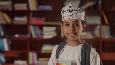 النظر الى الكاميرا بايماءات السرور والسعاده، التعليم فى سلطنة عمان ، الاستمتاع بقراءة الكتب المدرسية، ولد عربي خليجي عماني مبتسم يرتدي الدشداشه والكمه والحقيبة يقف ويمسك الكتاب بيده فى المكتبه المدرسيه