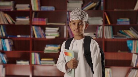 Enjoying reading school books, education in the Sultanate of Oman, a smiling Arab Gulf Omani boy wearing a dishdasha and kumma, standing and holding a book in his hand in the school library, looking at the camera with gestures of joy and happiness.