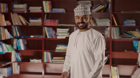 الاستمتاع بقراءة الكتب المدرسية، مدرس عربي خليجي عماني مبتسم يرتدي الدشداشه والكمه يقف بايدي متقاطعه فى المكتبه المدرسيه ، النظر الى الكاميرا بايماءات السرور والسعاده