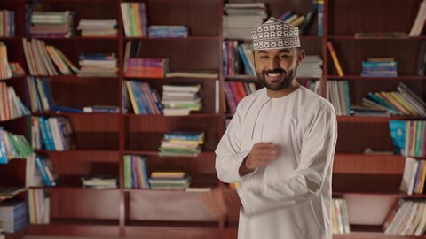 Looking at the camera with gestures of joy and happiness, enjoying reading school books, a smiling Arab Gulf Omani teacher wearing a dishdasha and kumma stands with crossed arms in the school library.