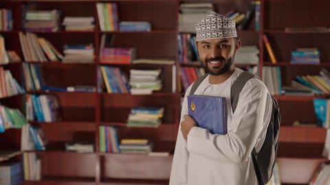مدرس عربي خليجي عماني مبتسم يرتدي الدشداشه والكمه والحقيبة يقف ويمسك الكتاب بيده فى المكتبه المدرسيه ،التعليم فى سلطنة عمان ، الاستمتاع بقراءة الكتب المدرسية،  النظر الى الكاميرا بايماءات السرور والسعاده