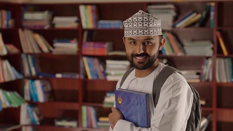 التعليم فى سلطنة عمان ، الاستمتاع بقراءة الكتب المدرسية، مدرس عربي خليجي عماني مبتسم يرتدي الدشداشه والكمه والحقيبة يقف ويمسك الكتاب بيده فى المكتبه المدرسيه ، النظر الى الكاميرا بايماءات السرور والسعاده