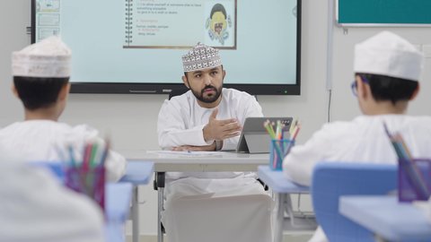 Developed curricula and e-learning, benefiting from modern technologies and technology in learning and studying, the evolution of teaching methods in schools in the Sultanate of Oman, two Saudi Gulf Arab students sitting in their seats with an Omani teacher using a tablet in front of them.