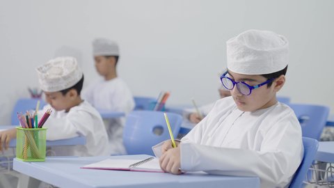 التعليم في سلطنة عمان ، تأدية الواجبات المدرسية، الإستعداد للعام الدراسي الجديد،العودة إلى المدارس، مجموعة من الطلاب العربيين الخليجيين العمانيين يرتدون الدشداشة والكمه يجلسون في مقاعدهم ويقومون بحل الواجب