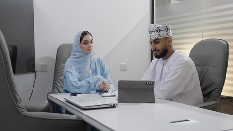 Data collection and problem-solving, a meeting and work discussion at the company headquarters, an Arab Gulf Omani businessman wearing a dishdasha and kumma working on a tablet with his colleague inside the office, developing and studying new projects in Omani companies, using modern technology, pointing to the tablet screen.
