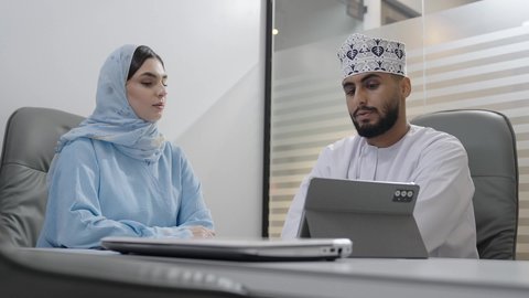 اجتماع ومناقشة عمل في مقر الشركة، رجل أعمال عربي خليجي عماني يرتدي الدشداشة و الكمة يقوم بالعمل على جهاز التابلت مع  زميلته داخل المكتب، تطوير ودراسة المشاريع الجديدة في الشركات العمانية، استخدام جهاز تقني حديث، جمع البيانات وحل المشكلات، الاشارة الى شاشة الجهاز اللوحي