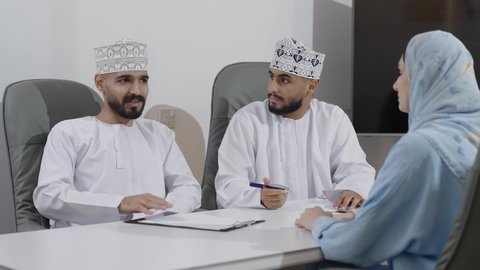 الحوار و النقاش بين الزملاء، تبادل الخبرات والمعلومات لتحقيق الاهداف, موظفان عربيان خليجيان عمانيان  يرتديان الدشداشة و الكمة يجلسان على مكتبهم داخل مقر الشركة برفقة زميلتهم، كتابة و تدوين المحلاظات ، بيئة العمل المريحة في الشركات العمانية