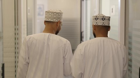 بيئة العمل المريحة في الشركات العمانية، مناقشة الخطط وشرح مهام الاعمال،تصوير من الخلف لزملاء عمل عربيان اماراتيون خليجيان يرتديان االدشداشة و الكمة  يقومان بالمشي داخل مقر الشركة، الاتفاق على المشاريع التجارية