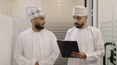 تبادل الآراء بين الزملاء، كتابة وتدوين ملاحظات لتحقيق الاستراتيجيات والغايات، تصوير مقرب  لرجل عربي خليجي عماني يرتدي الدشداشة و الكمة يسير داخل الشركه برفقة زميله يقوم بالشرح عن مشروع ، الاشارة الي شيء ما