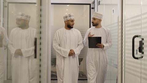 كتابة وتدوين ملاحظات لتحقيق الاستراتيجيات والغايات، تبادل الآراء بين الزملاء، رجل عربي خليجي عماني يرتدي الدشداشة و الكمة يسير داخل الشركه برفقة زميله يقوم بالشرح عن مشروع ، الاشارة الي شيء ما