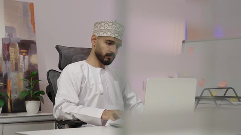 دمج العمل مع التقنية والتكنولوجيا، ايماءات الاندماج و التركيز، النقر على لوحه المفاتيح ، إدارة شؤون الأعمال وتنظيمها عبر الاجهزة التقنية، مهن ووظائف ادارية مكتبية، رجل عربي خليجي عماني يرتدي الدشداشة و الكمة يقوم بالعمل على الحاسوب المحمول داخل المكتب