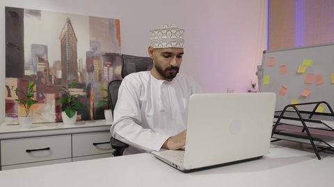 النقر على لوحه المفاتيح ، إدارة شؤون الأعمال وتنظيمها عبر الاجهزة التقنية، مهن ووظائف ادارية مكتبية، دمج العمل مع التقنية والتكنولوجيا، رجل عربي خليجي عماني يرتدي الدشداشة و الكمة يقوم ,بالعمل على الحاسوب المحمول داخل المكتب، ايماءات الاندماج و التركيز