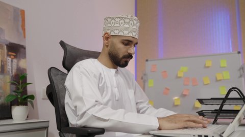 إدارة شؤون الأعمال وتنظيمها عبر الاجهزة التقنية، مهن ووظائف ادارية مكتبية، دمج العمل مع التقنية والتكنولوجيا، تصوير من الجانب لرجل عربي خليجي عماني يرتدي الدشداشة و الكمة يقوم ,بالعمل على الحاسوب المحمول داخل المكتب، النقر على لوحه المفاتيح ، ايماءات الاندماج و التركيز