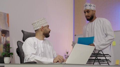 Studying statistical data sheets, developing projects and investments, cooperation and understanding among colleagues, and setting strategic plans, a photo of an Arab Gulf Omani man wearing a dishdasha and kumma sitting in front of a laptop writing and taking notes.