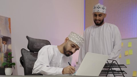 روح الفريق الواحد والعمل الجماعي، تبادل الخبرات والمعلومات لتحقيق الاهداف، التعاون والتفاهم بين الزملاء ووضع الخطط الاستراتيجية،ايماءات السعادة و السرور، تصوير لرجل عربي خليجي عماني يرتدي الدشداشة و الكمة يجلس امام الحاسب المحمول يقوم  بكتابة و تدوين الملاحظات