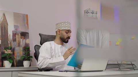 تبادل الخبرات والمعلومات لتحقيق الاهداف، التعاون والتفاهم بين الزملاء ووضع الخطط الاستراتيجية، عقد اجتماع داخل مقر الشركة، تصوير لرجل عربي خليجي عماني يرتدي الدشداشة و الكمة يجلس امام الحاسب المحمول يمسك بيديه ملف ازرق ، روح الفريق الواحد والعمل الجماعي
