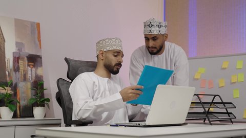 التعاون والتفاهم بين الزملاء ووضع الخطط الاستراتيجية، عقد اجتماع داخل مقر الشركة، تصوير لرجل عربي خليجي عماني يرتدي الدشداشة و الكمة يجلس امام الحاسب المحمول يمسك بيديه ملف ازرق ، تبادل الخبرات والمعلومات لتحقيق الاهداف، روح الفريق الواحد والعمل الجماعي